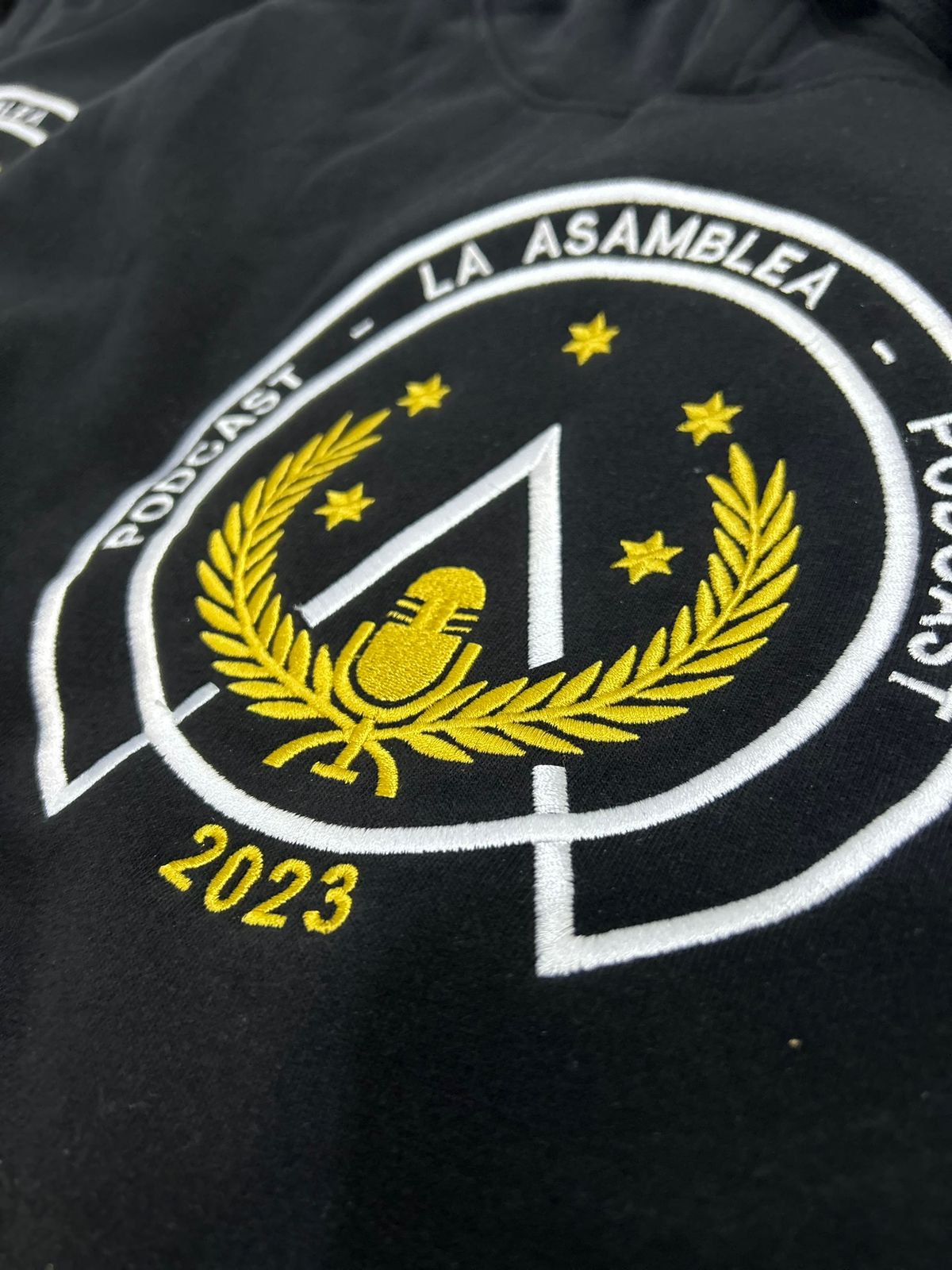 Hoodie negro con escudo bordado de Asamblea Podcast 2023 en hilos dorado y blanco